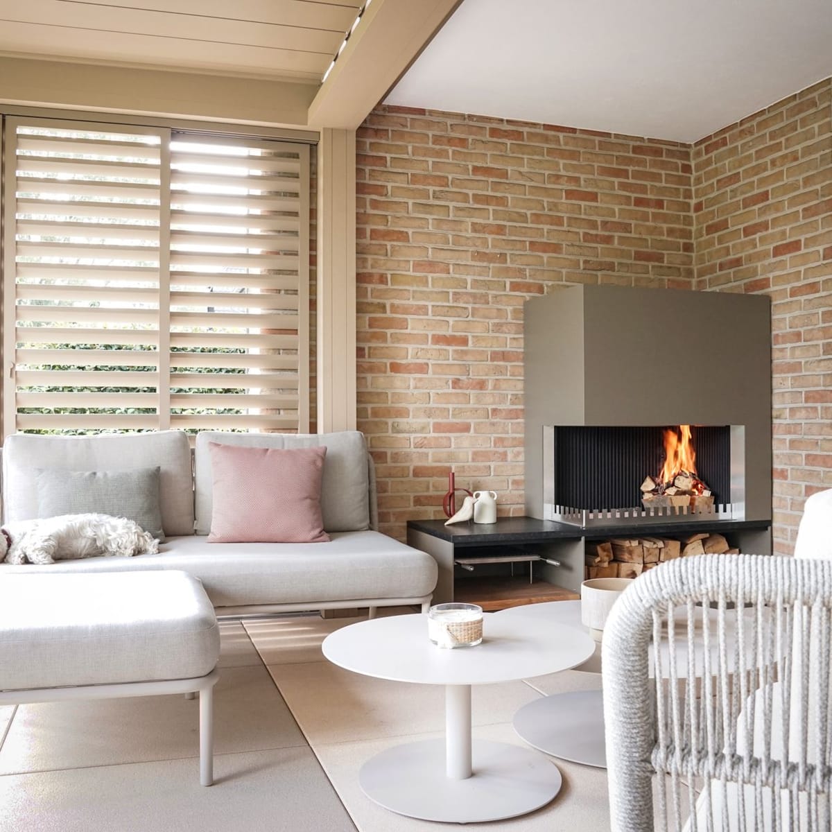 Covered lounge with louvered roof, sofas, round tables and a wood fire in a steel fireplace unit, dog on the sofa