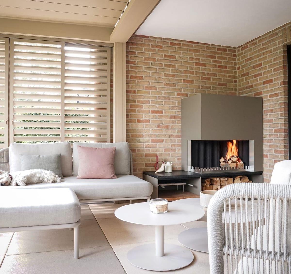 Covered lounge with louvered roof, sofas, round tables and a wood fire in a steel fireplace unit, dog on the sofa