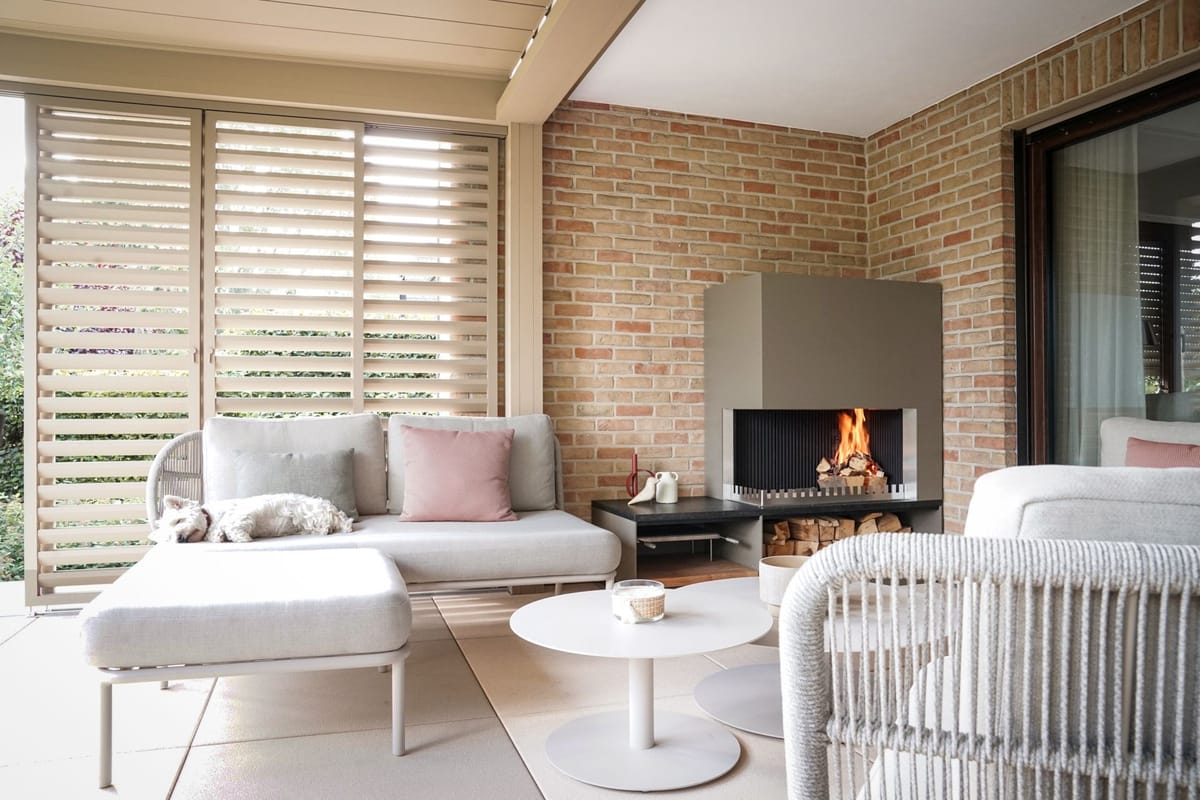 Covered lounge with louvered roof, sofas, round tables and a wood fire in a steel fireplace unit, dog on the sofa
