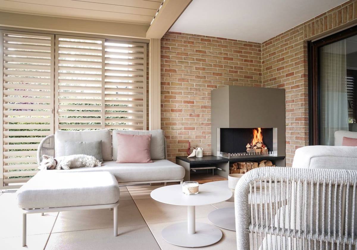 Covered lounge with louvered roof, sofas, round tables and a wood fire in a steel fireplace unit, dog on the sofa
