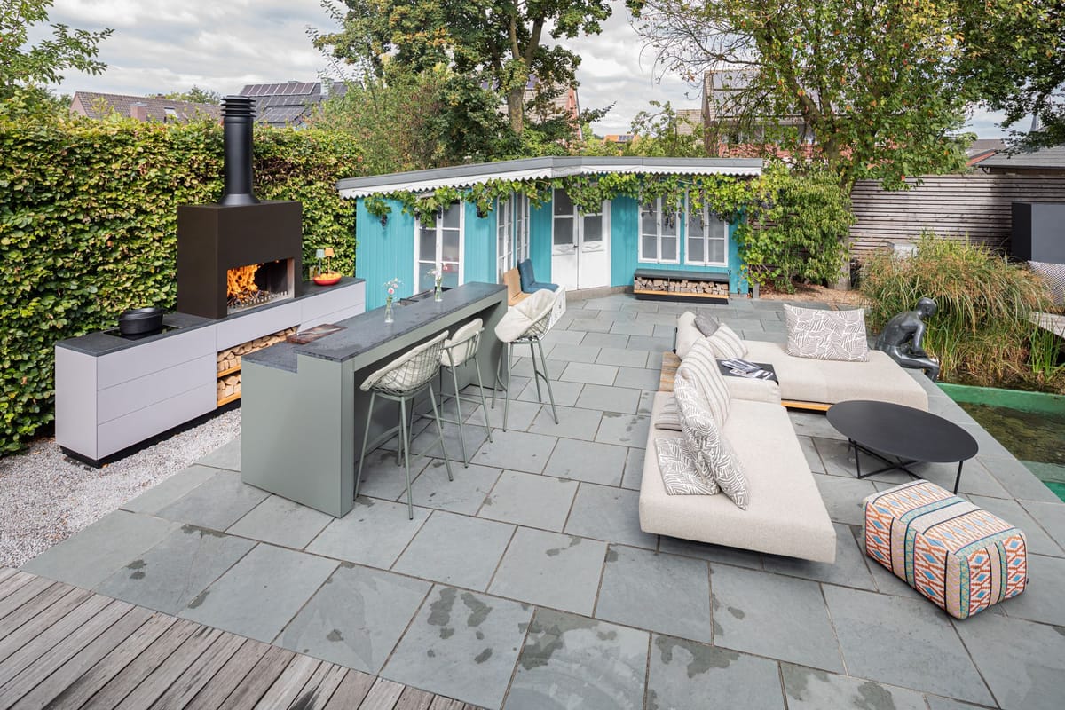 Garden patio with a wood fire under a black steel hood, light gray kitchen, green bar with stools and a lounge sofa
