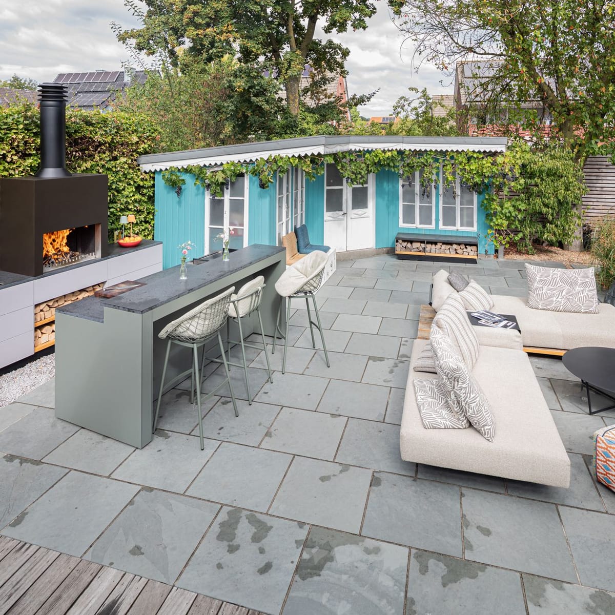 Garden patio with a wood fire under a black steel hood, light gray kitchen, green bar with stools and a lounge sofa