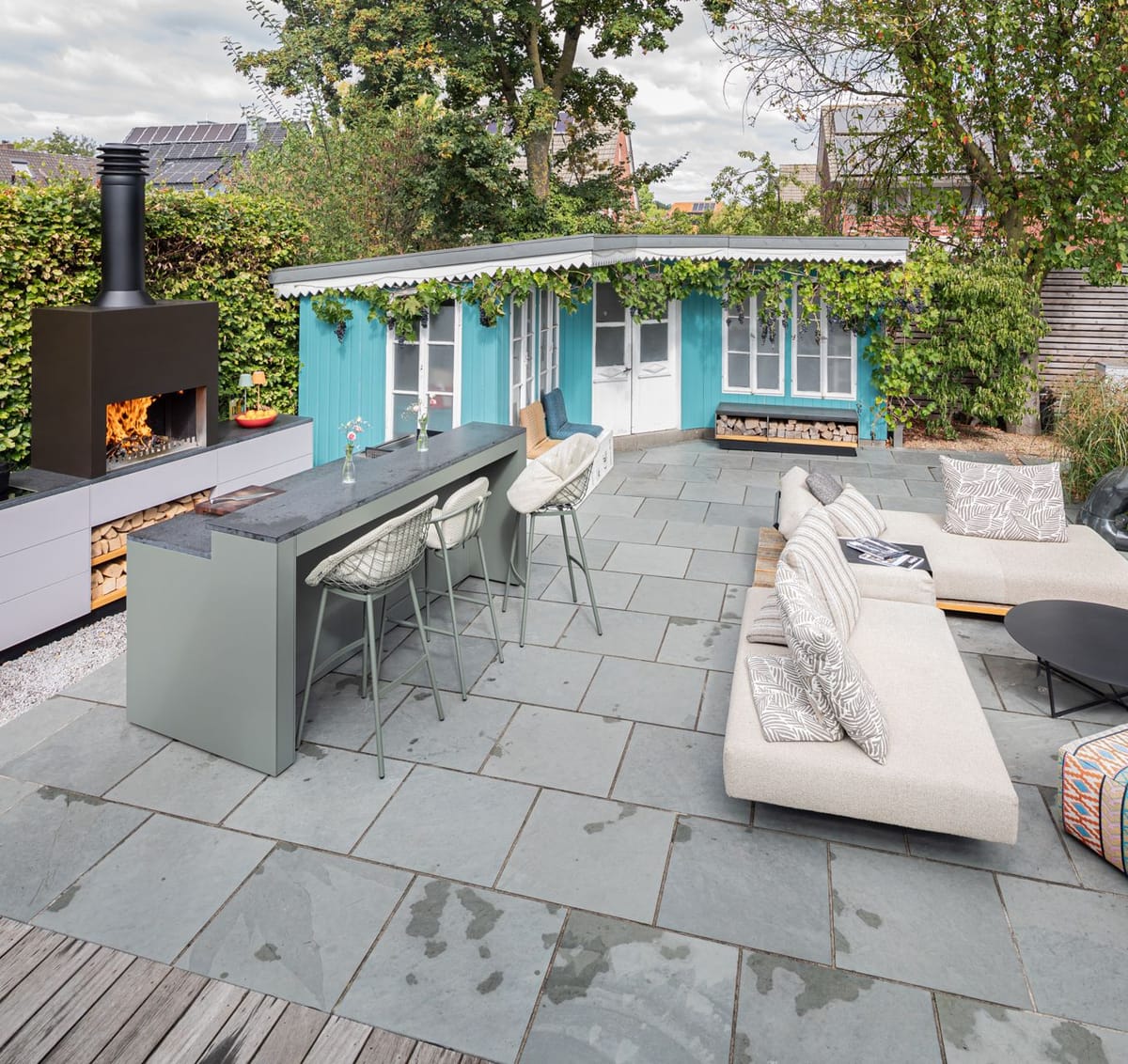 Garden patio with a wood fire under a black steel hood, light gray kitchen, green bar with stools and a lounge sofa
