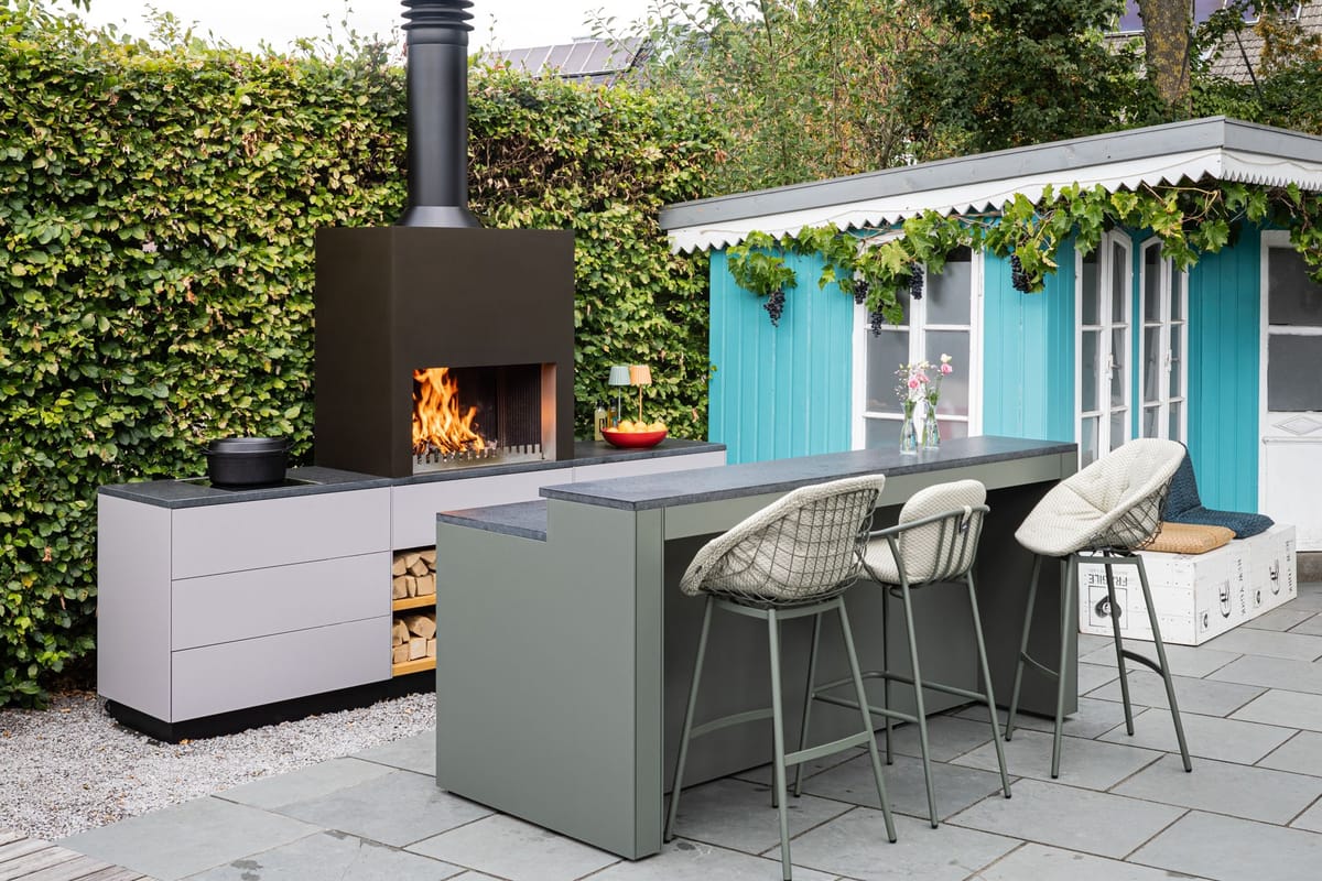 Wood fire under a black steel hood above a light gray kitchen with log storage, green bar counter with three stools