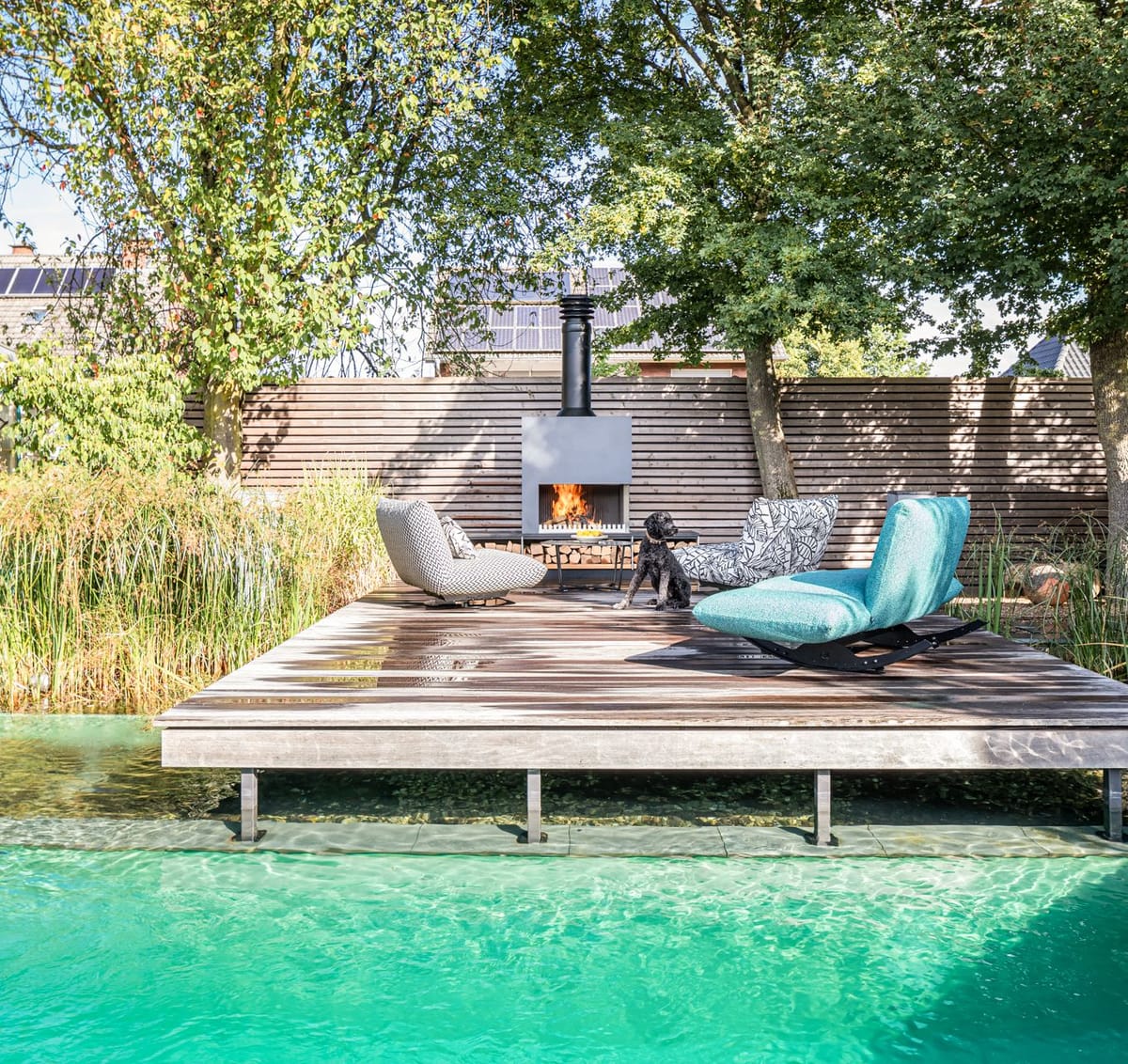 Wooden deck over a clear natural pool with lounge chairs around a freestanding wood-burning fireplace, dog on deck