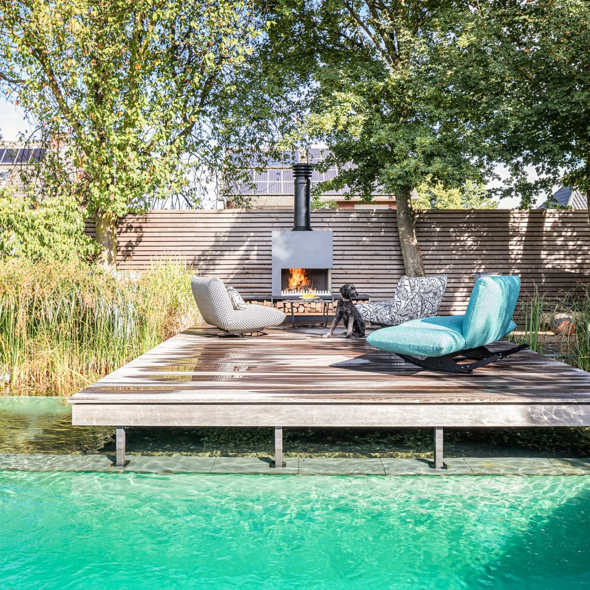 Wooden deck over a clear natural pool with lounge chairs around a freestanding wood-burning fireplace, dog on deck
