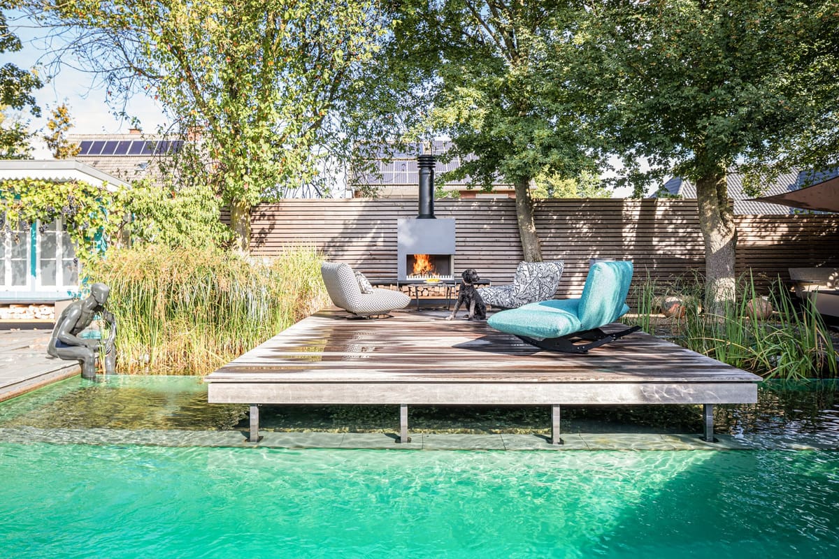 Wooden deck over a clear natural pool with lounge chairs around a freestanding wood-burning fireplace, dog on deck