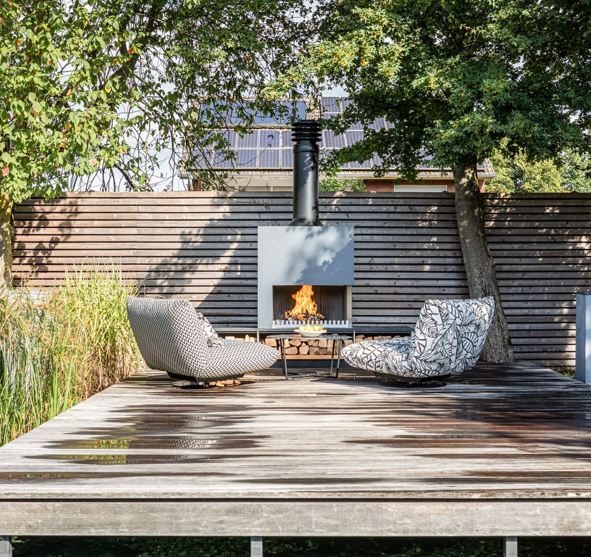Weathered wooden deck with two patterned lounge chairs facing a freestanding wood-burning fireplace with log storage