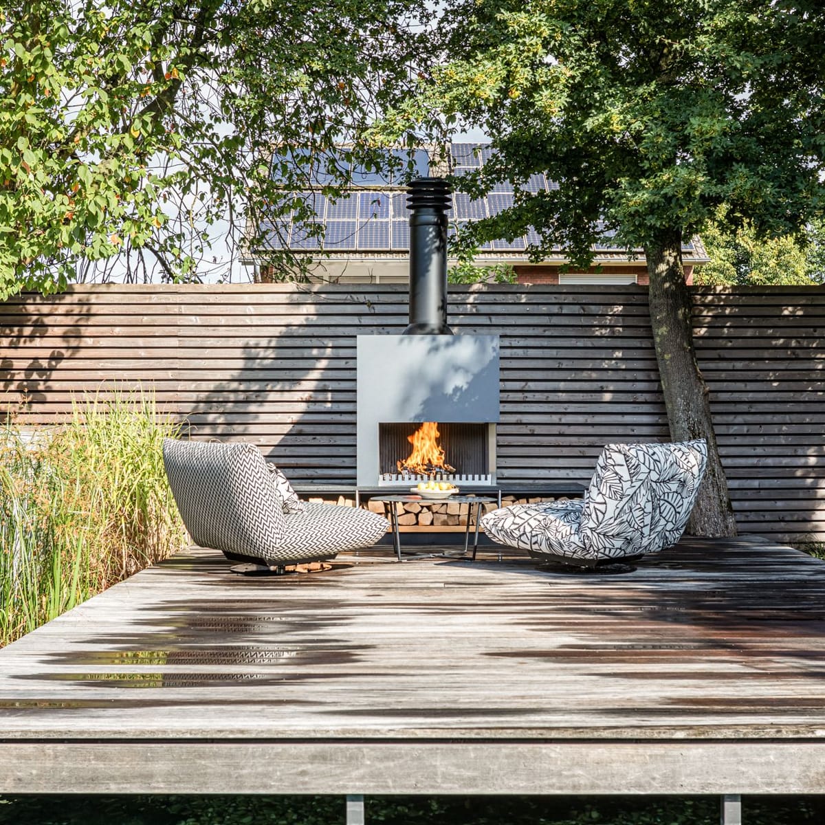 Weathered wooden deck with two patterned lounge chairs facing a freestanding wood-burning fireplace with log storage