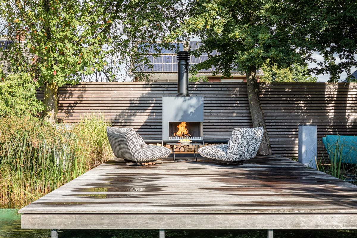 Weathered wooden deck with two patterned lounge chairs facing a freestanding wood-burning fireplace with log storage