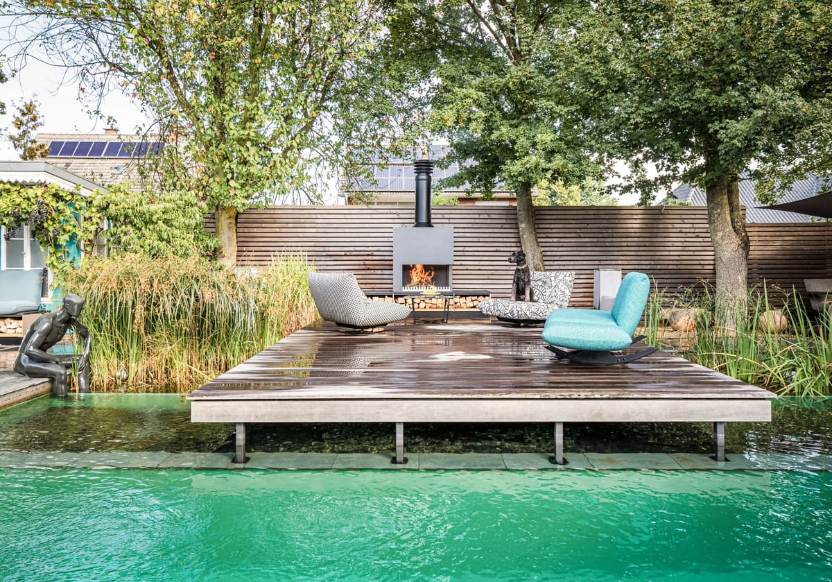 Natural pool with a wooden deck, lounge chairs and a freestanding wood-burning fireplace, dog on a chair, trees behind