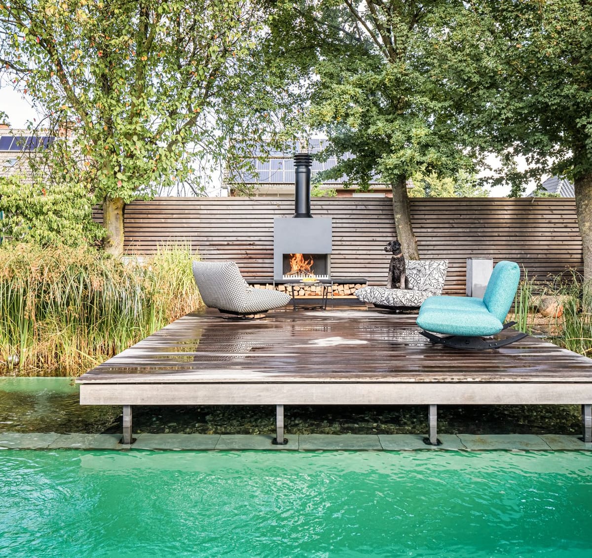Natural pool with a wooden deck, lounge chairs and a freestanding wood-burning fireplace, dog on a chair, trees behind