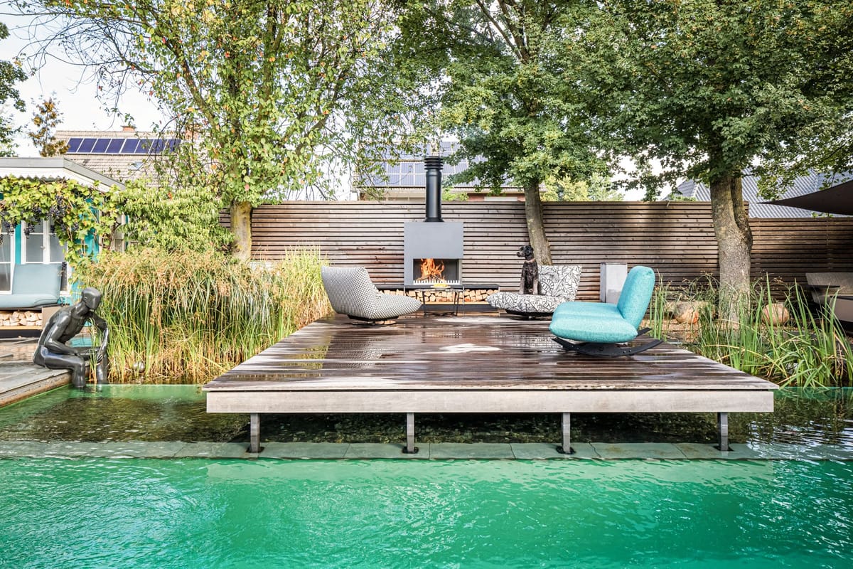 Natural pool with a wooden deck, lounge chairs and a freestanding wood-burning fireplace, dog on a chair, trees behind