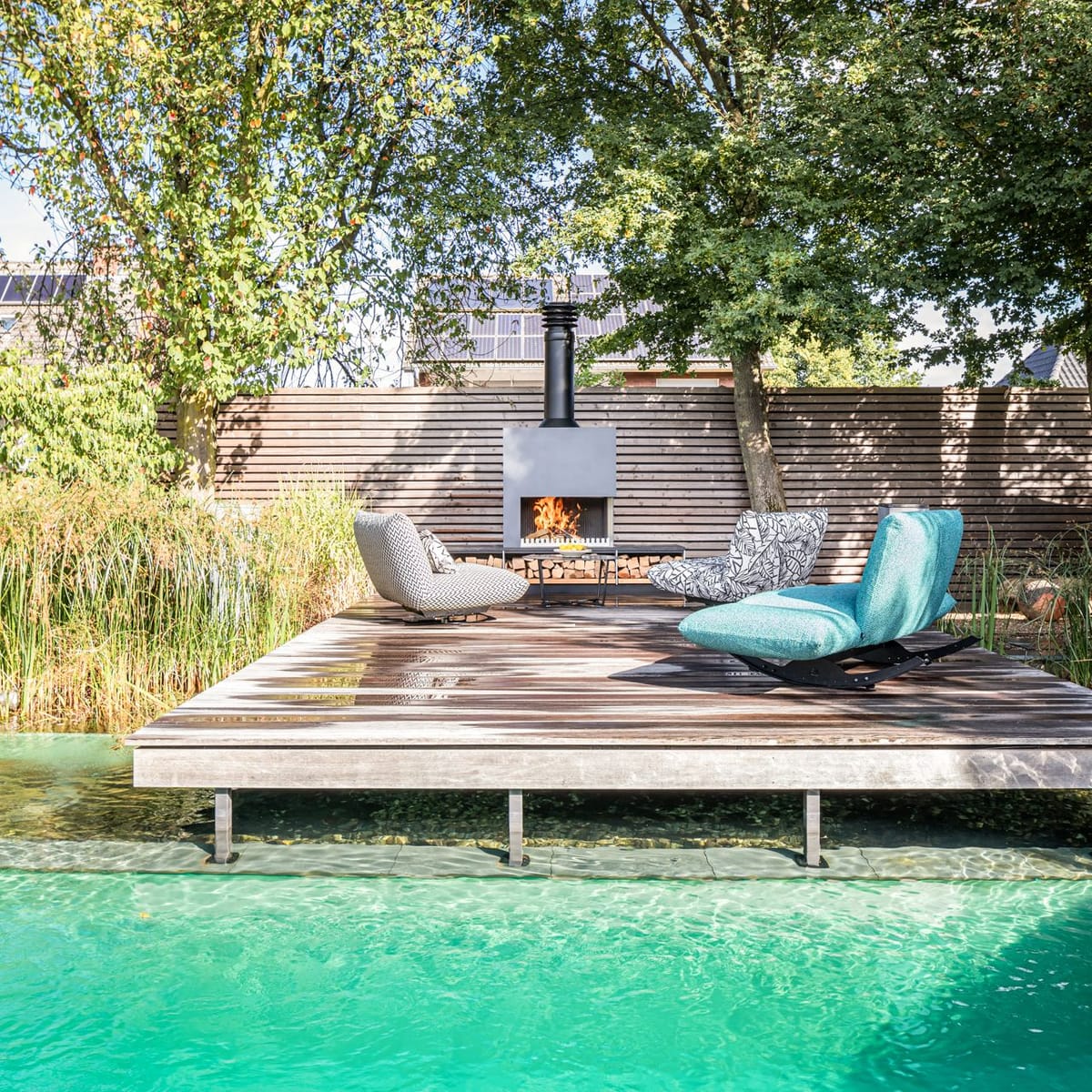 Natural pool with a floating wooden deck, lounge chairs and a freestanding wood-burning fireplace under tall trees
