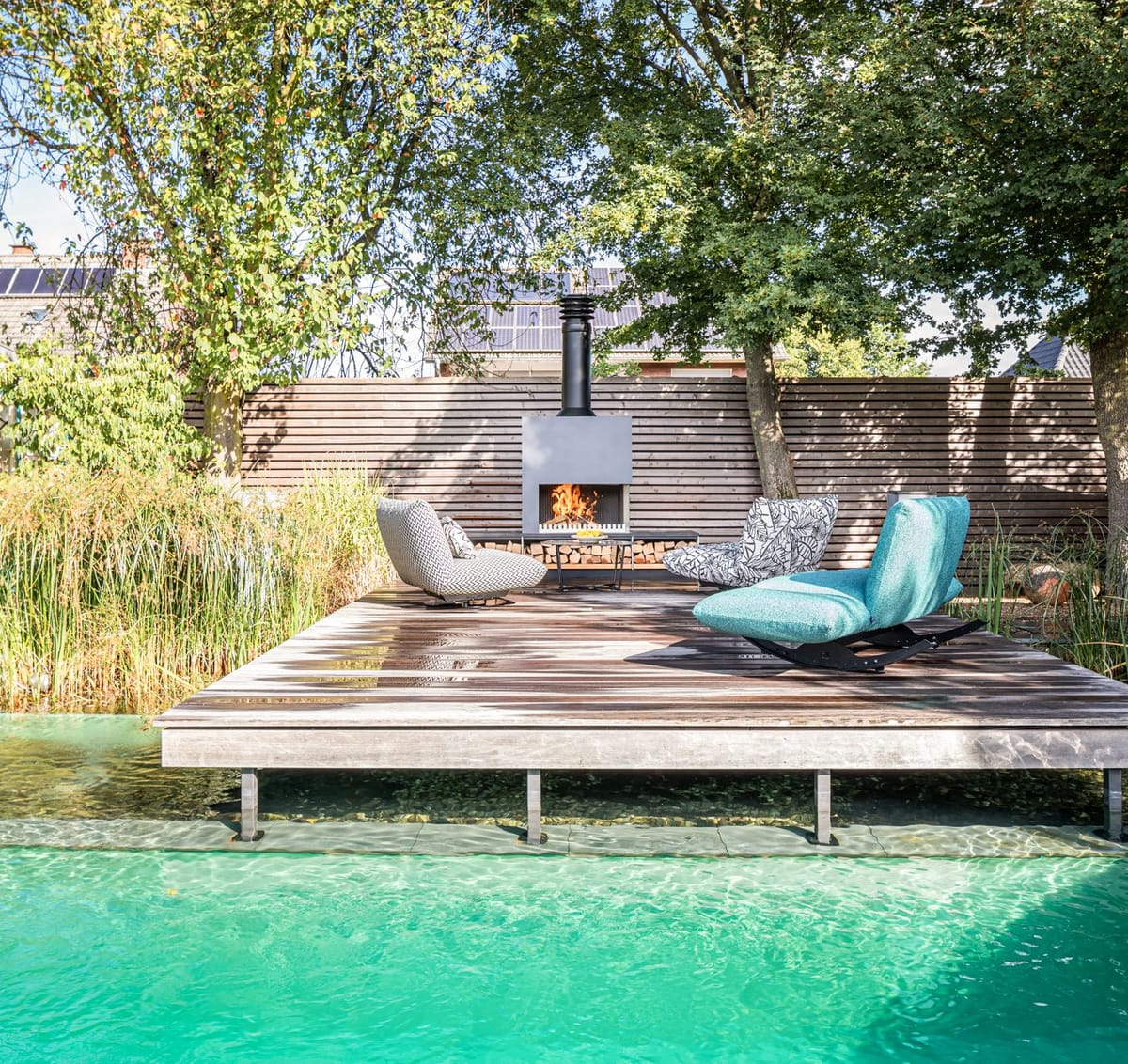 Natural pool with a floating wooden deck, lounge chairs and a freestanding wood-burning fireplace under tall trees