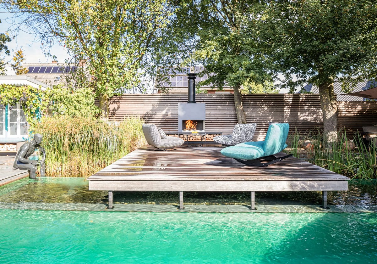 Natural pool with a floating wooden deck, lounge chairs and a freestanding wood-burning fireplace under tall trees