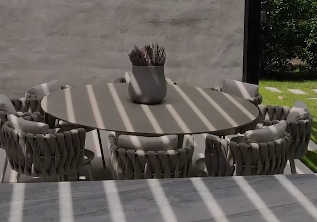 A round dining table under a louvered pergola, seen across the stone worktop of a FREILUFT kitchen run