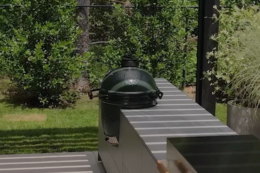 A Big Green Egg set into the stone worktop of a FREILUFT kitchen run, next to a built-in grill, under a louvered pergola