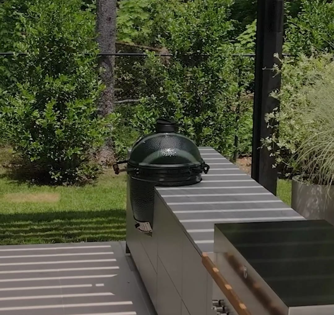 A Big Green Egg set into the stone worktop of a FREILUFT kitchen run, next to a built-in grill, under a louvered pergola