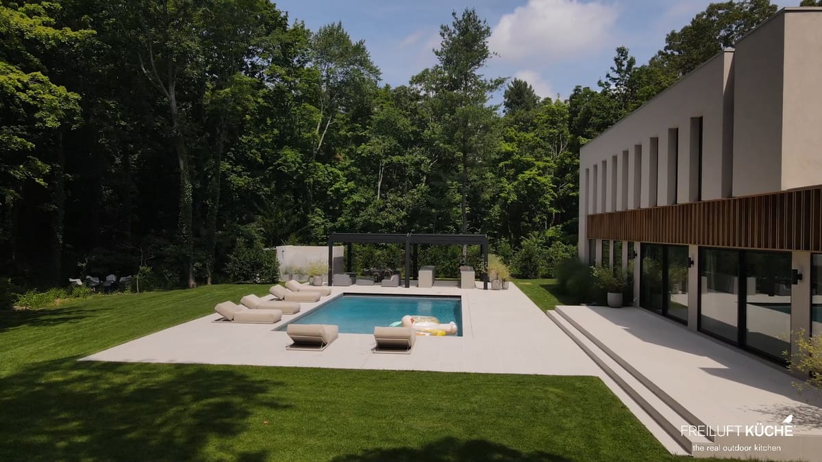 A modern house on Long Island, New York, with a lawn, a pool and a black pergola with a FREILUFT kitchen at the far end of the pool