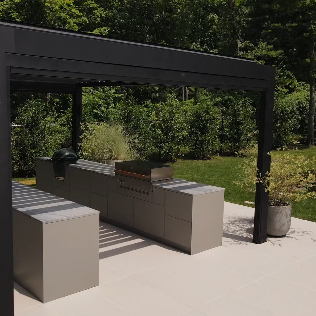 Two long gray FREILUFT kitchen runs under a black pergola in a garden on Long Island, New York, with a built-in grill and a Big Green Egg