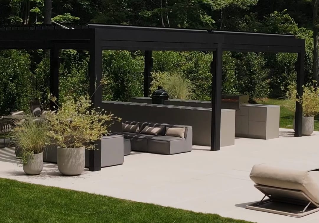 A black louvered pergola with a lounge and FREILUFT kitchen runs, seen from the lawn of a garden on Long Island, New York
