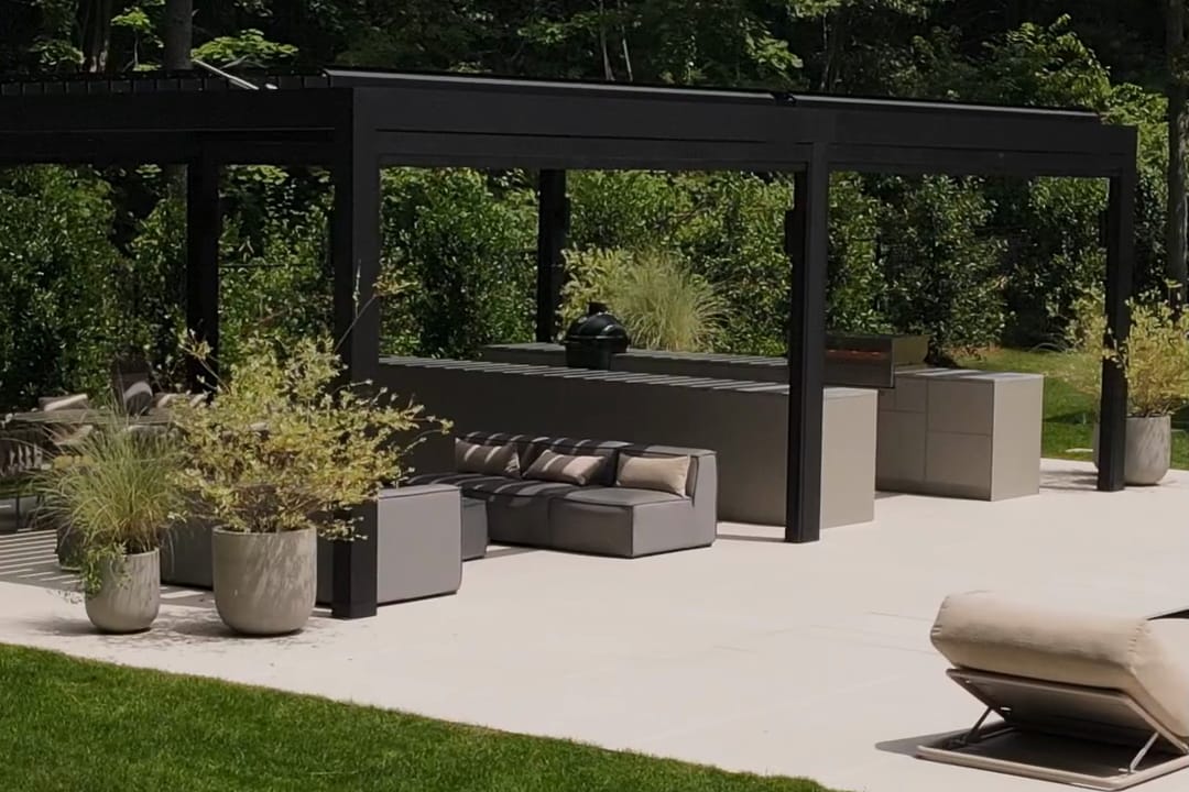 A black louvered pergola with a lounge and FREILUFT kitchen runs, seen from the lawn of a garden on Long Island, New York