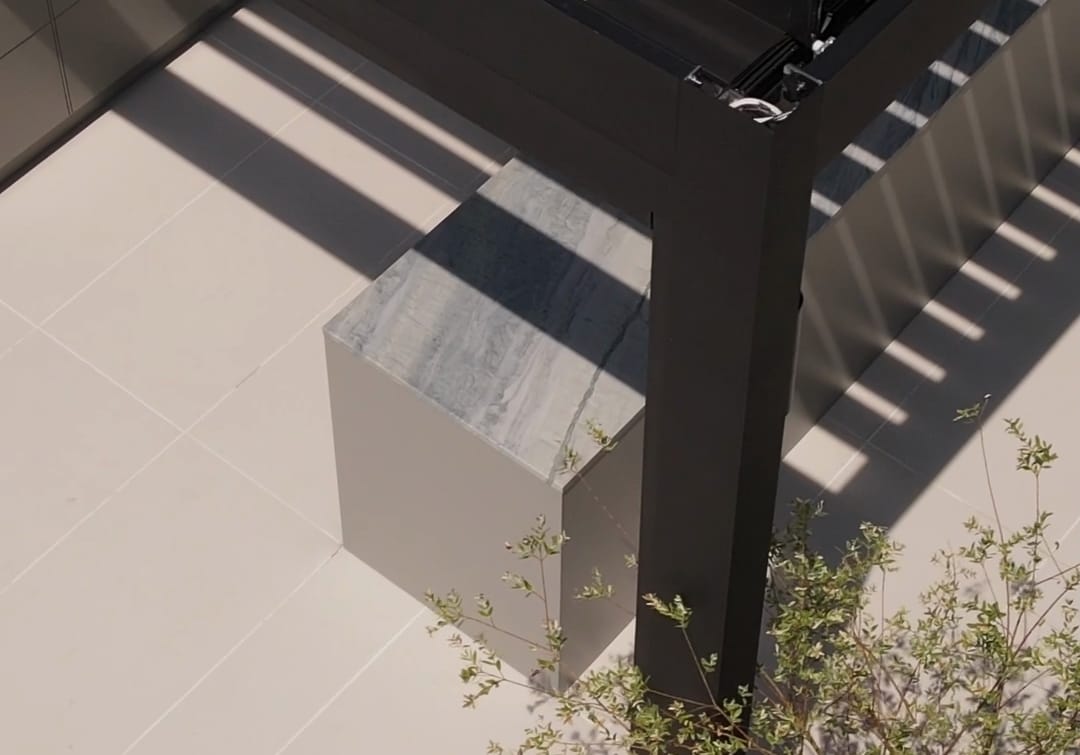 The corner of a FREILUFT cabinet with a gray-veined stone worktop beside the black post of a louvered pergola