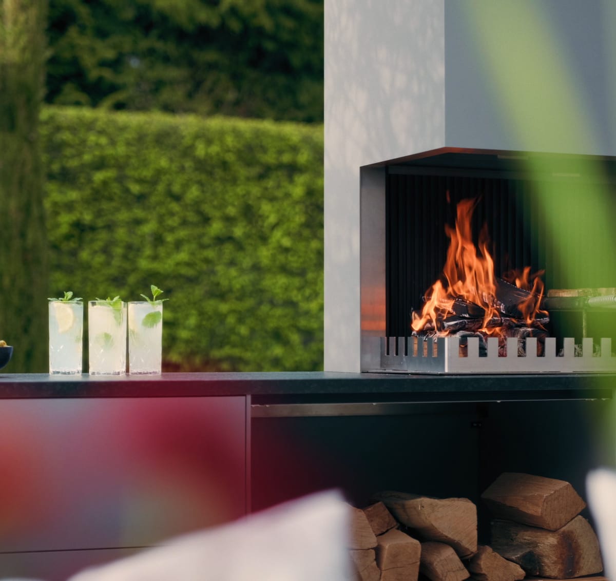 A light gray FREILUFT grill-and-fire element with a wood fire and open log storage on a garden terrace, green hedge and glass drinks in the foreground