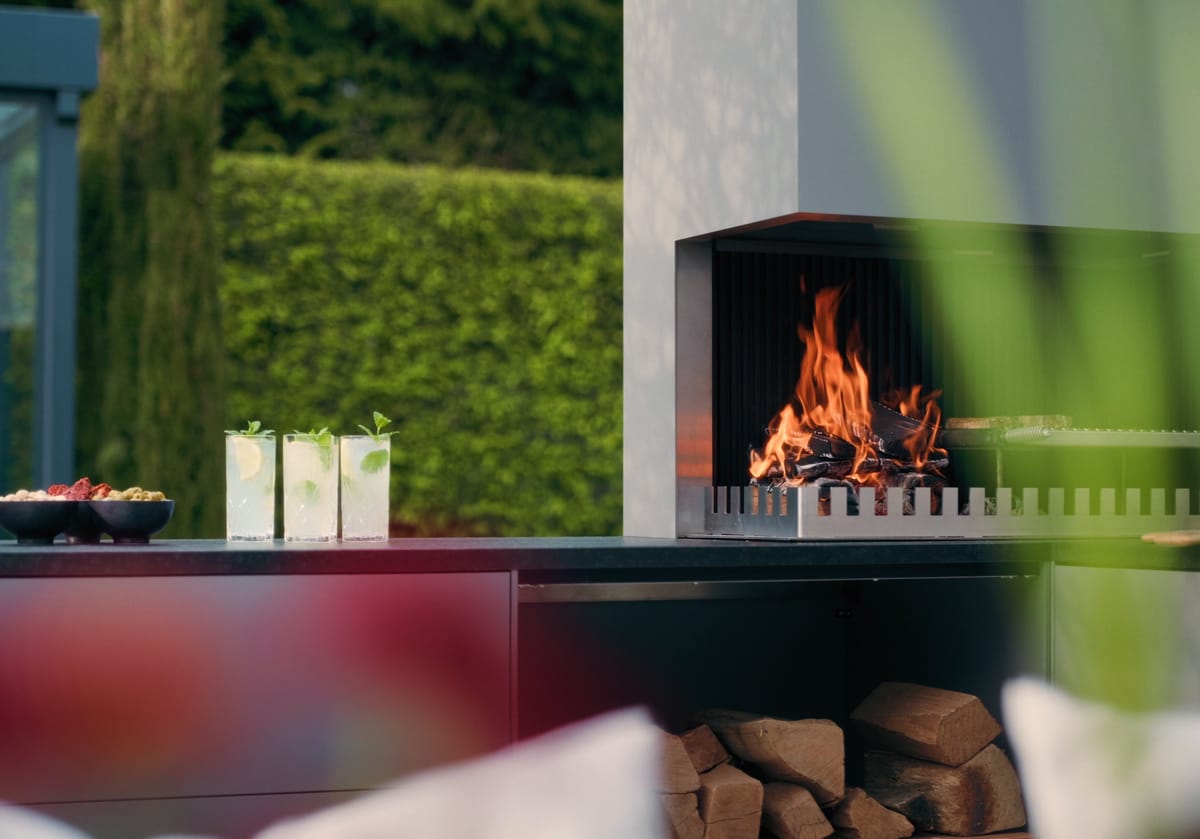 A light gray FREILUFT grill-and-fire element with a wood fire and open log storage on a garden terrace, green hedge and glass drinks in the foreground