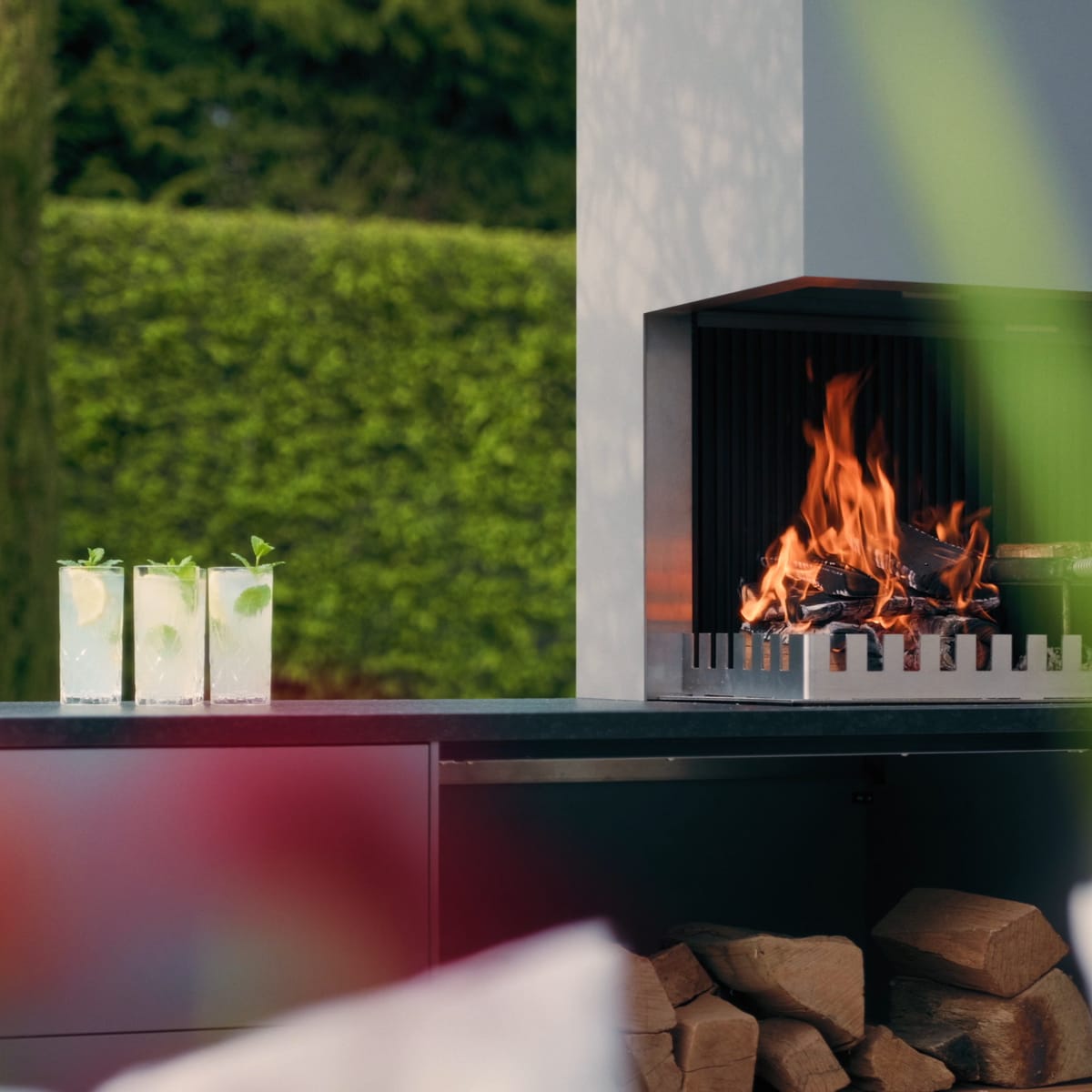 A light gray FREILUFT grill-and-fire element with a wood fire and open log storage on a garden terrace, green hedge and glass drinks in the foreground