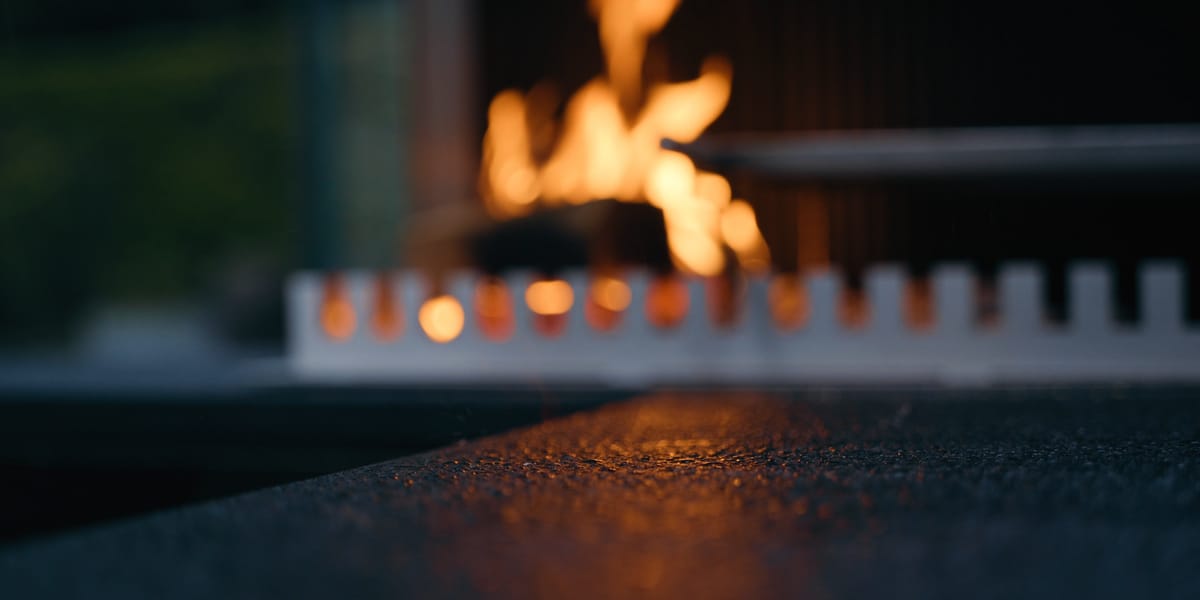 A wood fire behind the notched steel edge of a grill-and-fire element, seen low across a dark stone countertop at dusk