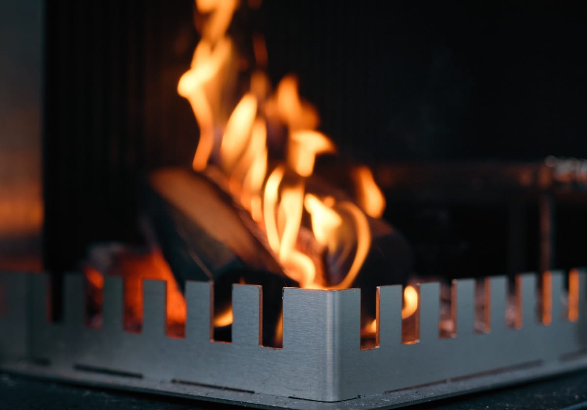 Close-up of a wood fire burning behind the notched steel edge of a FREILUFT grill-and-fire element at dusk