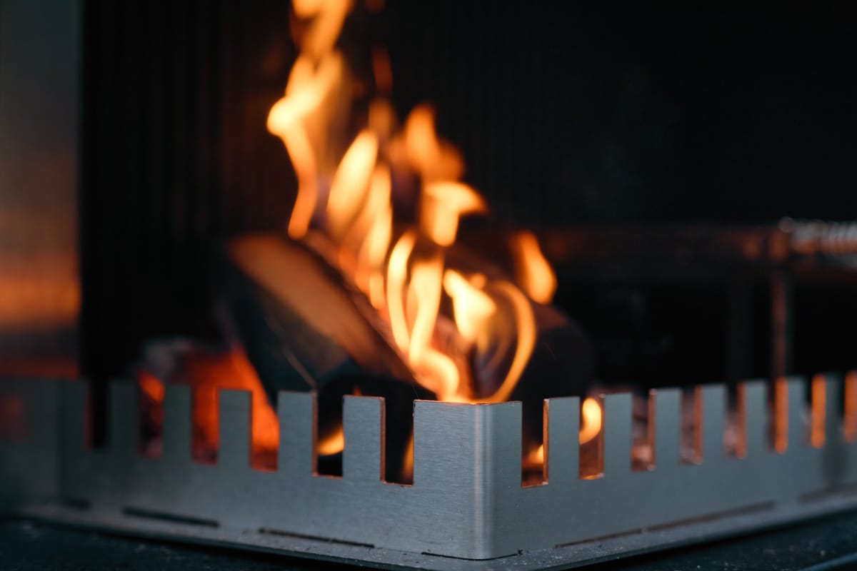 Close-up of a wood fire burning behind the notched steel edge of a FREILUFT grill-and-fire element at dusk