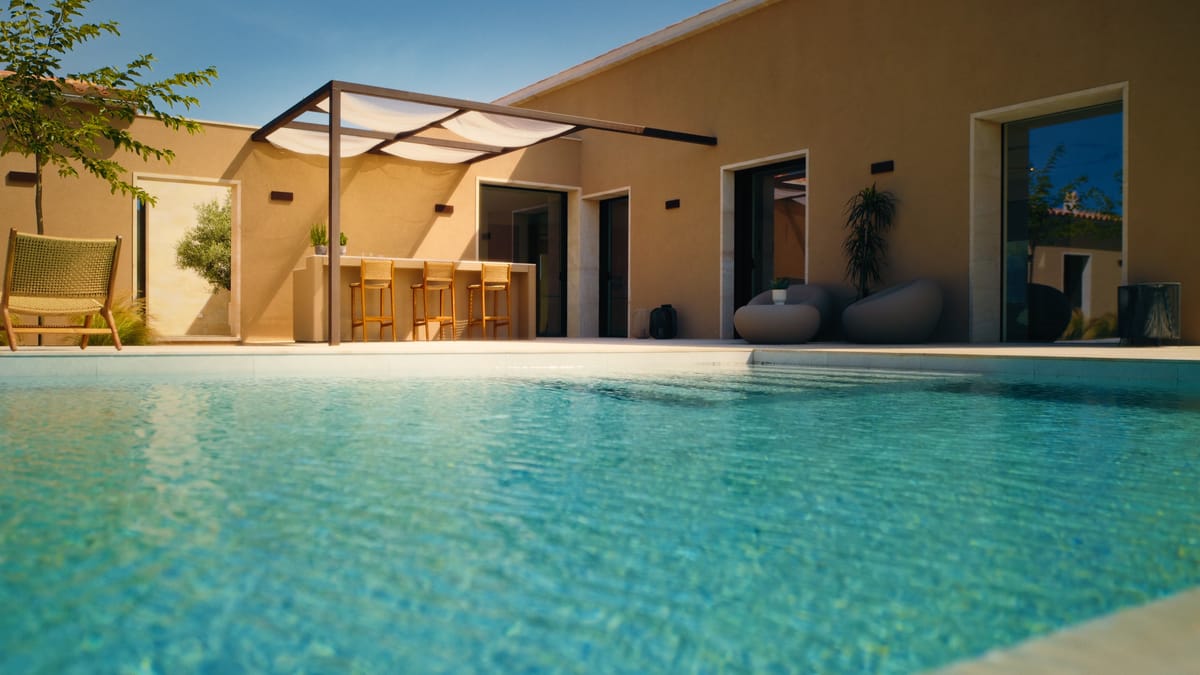 A turquoise pool in front of a warm stucco house, an outdoor bar with three stools under a pergola with sail shades