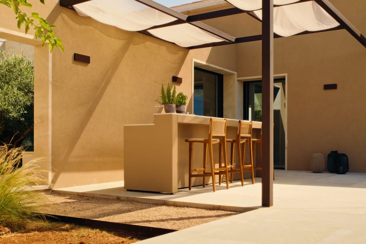 A sand-colored FREILUFT bar counter with three wooden stools under a pergola with a sunshade, beside a sand-colored house