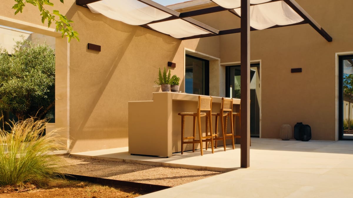 A sand-colored FREILUFT bar counter with three wooden stools under a pergola with a sunshade, beside a sand-colored house