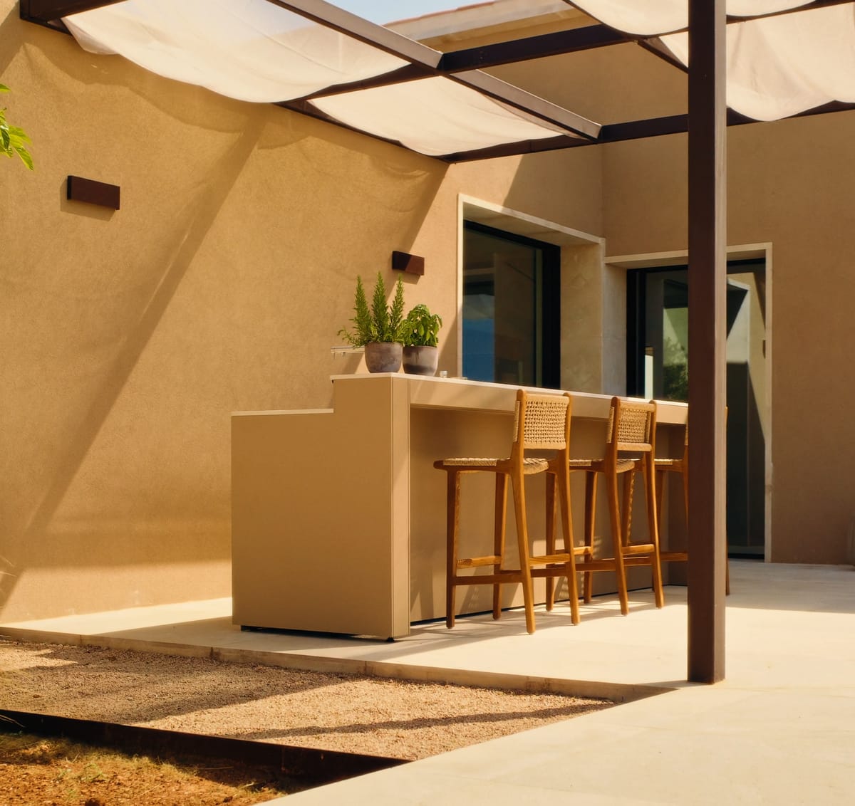 A sand-colored FREILUFT bar counter with three wooden stools under a pergola with a sunshade, beside a sand-colored house