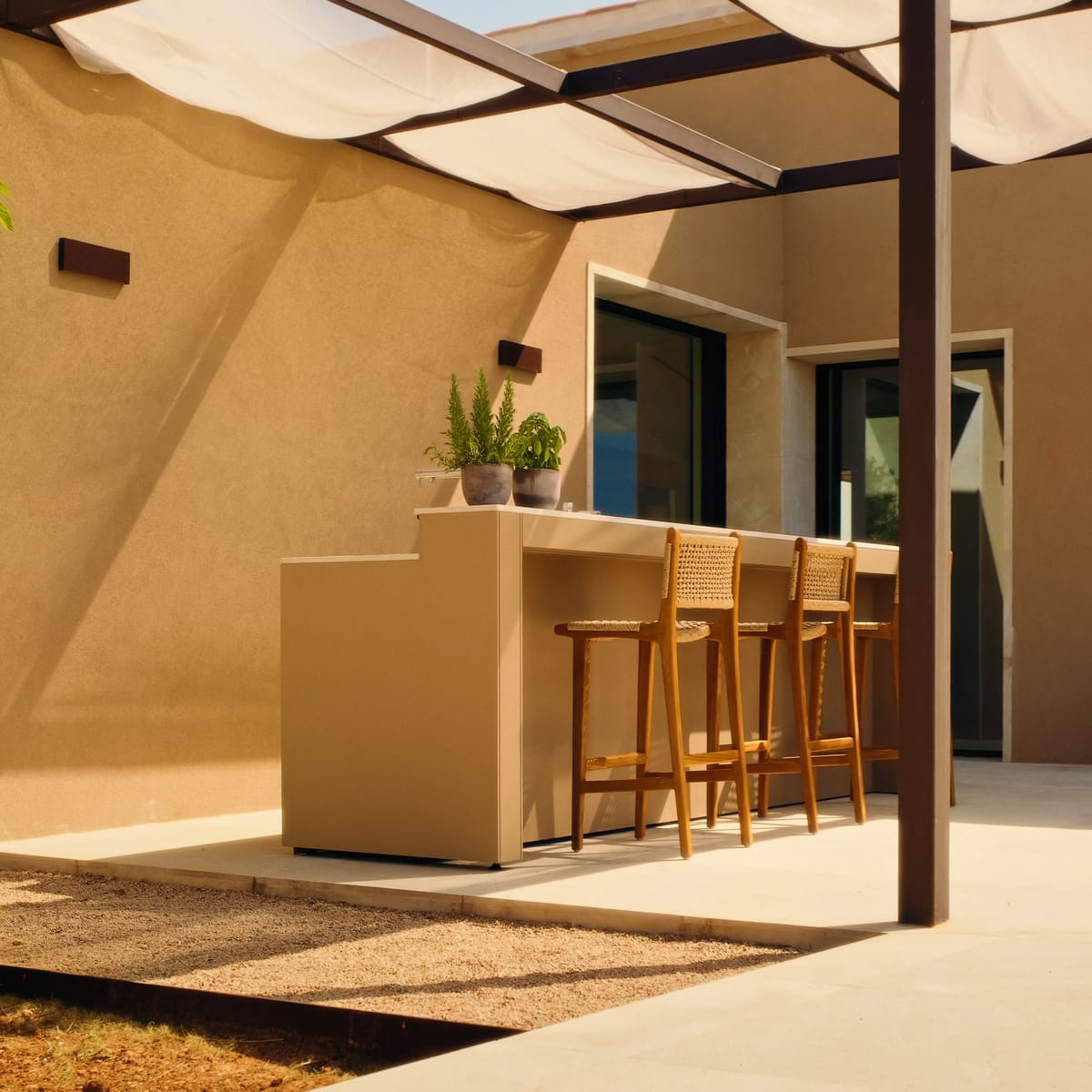 A sand-colored FREILUFT bar counter with three wooden stools under a pergola with a sunshade, beside a sand-colored house