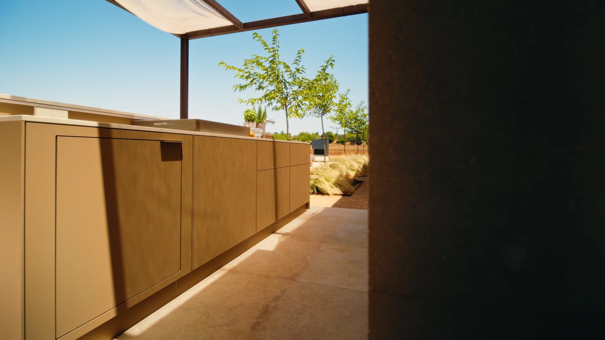 A long sand-colored FREILUFT kitchen run with handleless fronts under a pergola in bright sunlight