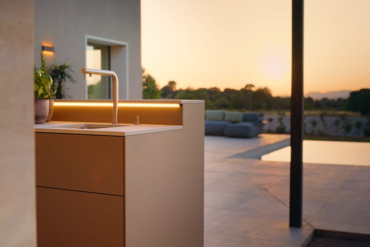 A sand-colored kitchen unit with a sink and a warm LED light strip, a terrace and the evening sky behind it