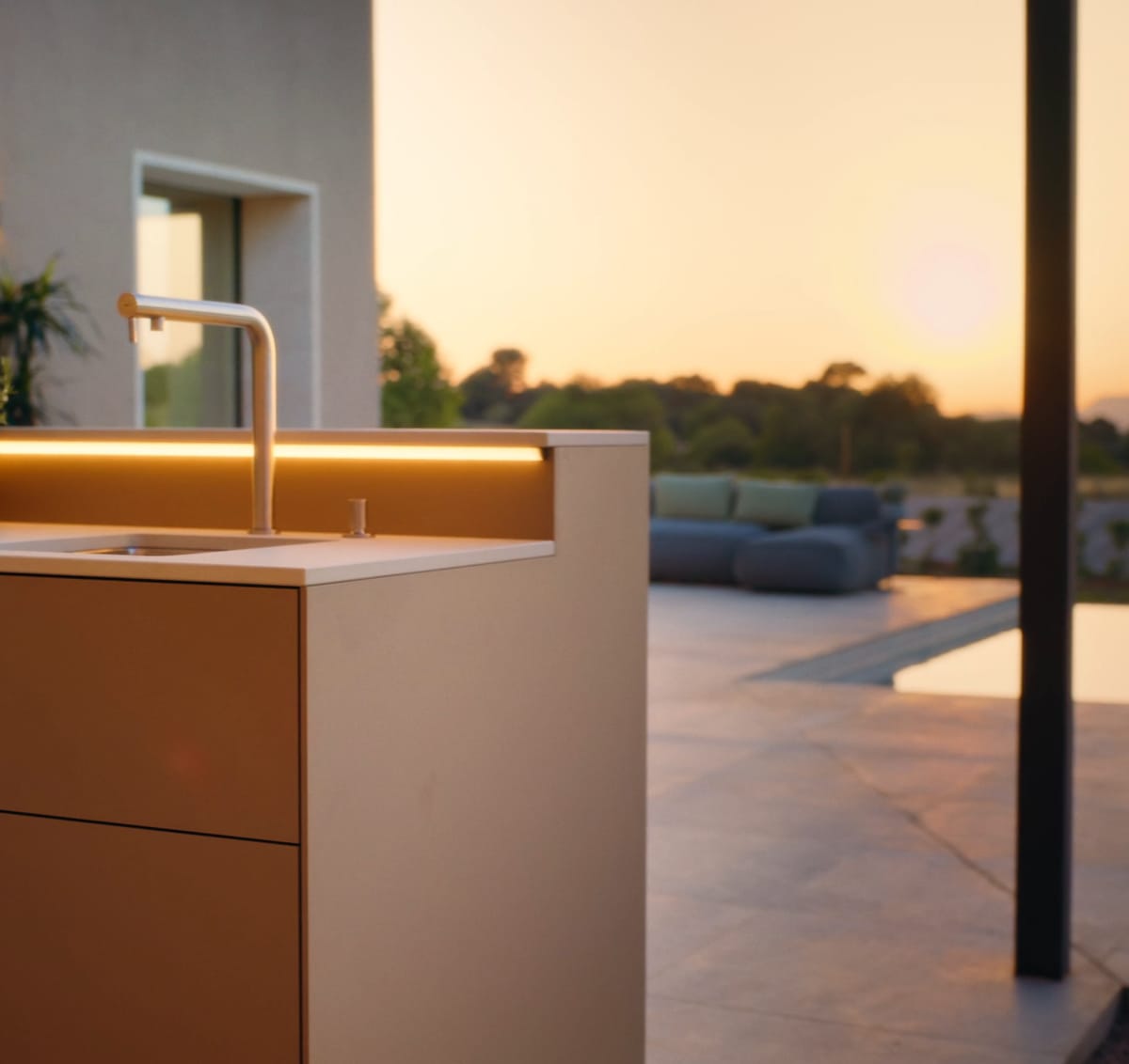 A sand-colored kitchen unit with a sink and a warm LED light strip, a terrace and the evening sky behind it