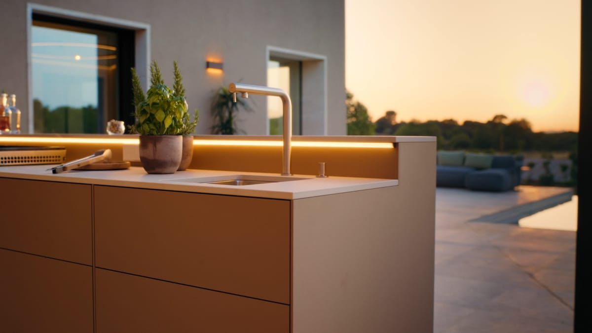 A FREILUFT kitchen run with a sink, herbs and a warm LED strip in front of a house, a pool and the setting sun behind it
