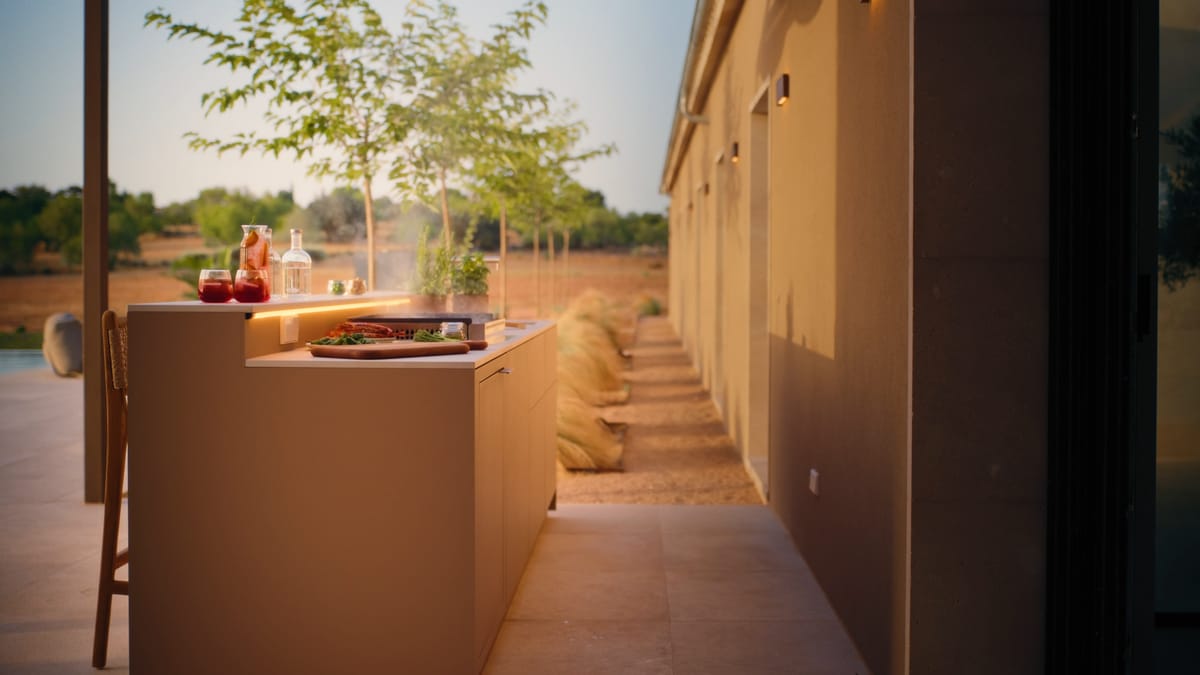 A sand-colored FREILUFT kitchen run along a stucco wall in evening light, drinks, herbs and a cutting board on the counter under a warm LED strip