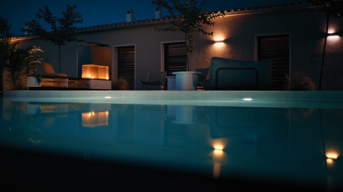 A pool at night with an underwater light, a house with warm wall lights and a glowing outdoor fire on the terrace behind it