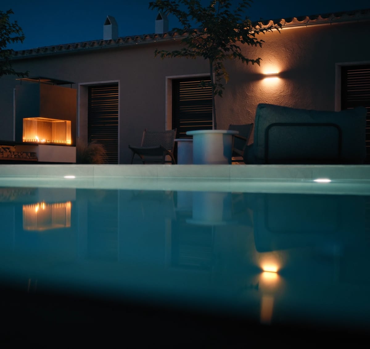 A pool at night with an underwater light, a house with warm wall lights and a glowing outdoor fire on the terrace behind it