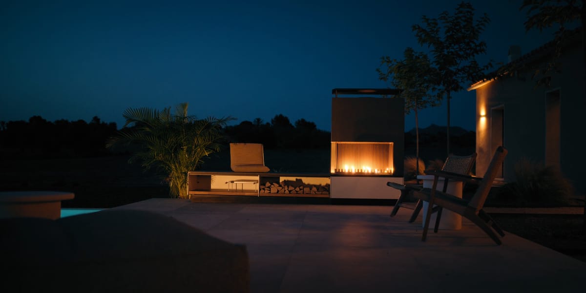 A fire burning in a light outdoor fireplace on a terrace beside a pool at night, lounge chairs in front of it