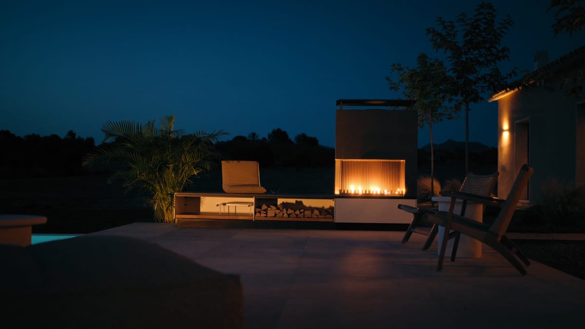 A fire burning in a light outdoor fireplace on a terrace beside a pool at night, lounge chairs in front of it