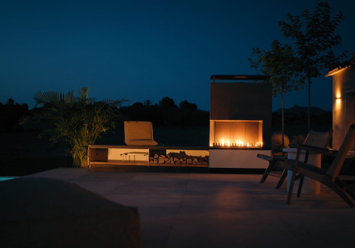 A fire burning in a light outdoor fireplace on a terrace beside a pool at night, lounge chairs in front of it