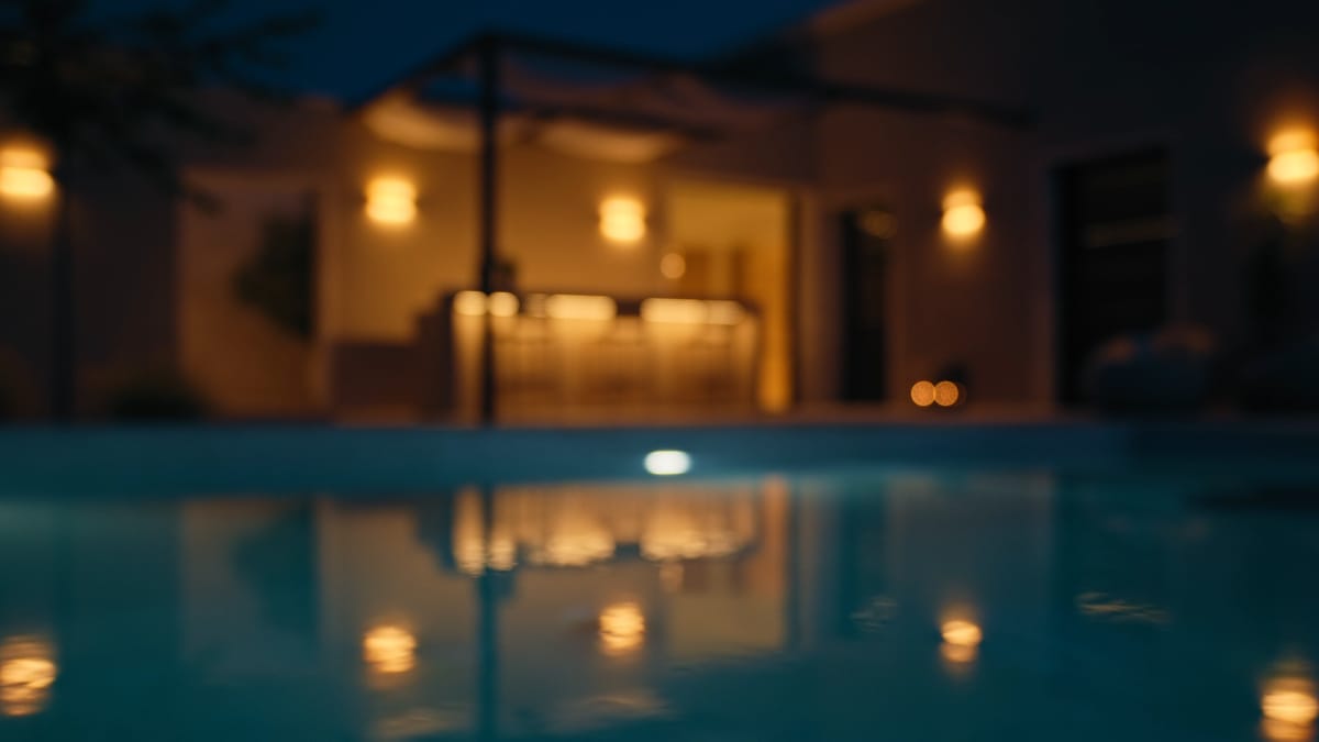 A pool at night reflecting the warm lights of a house and an outdoor kitchen under a pergola